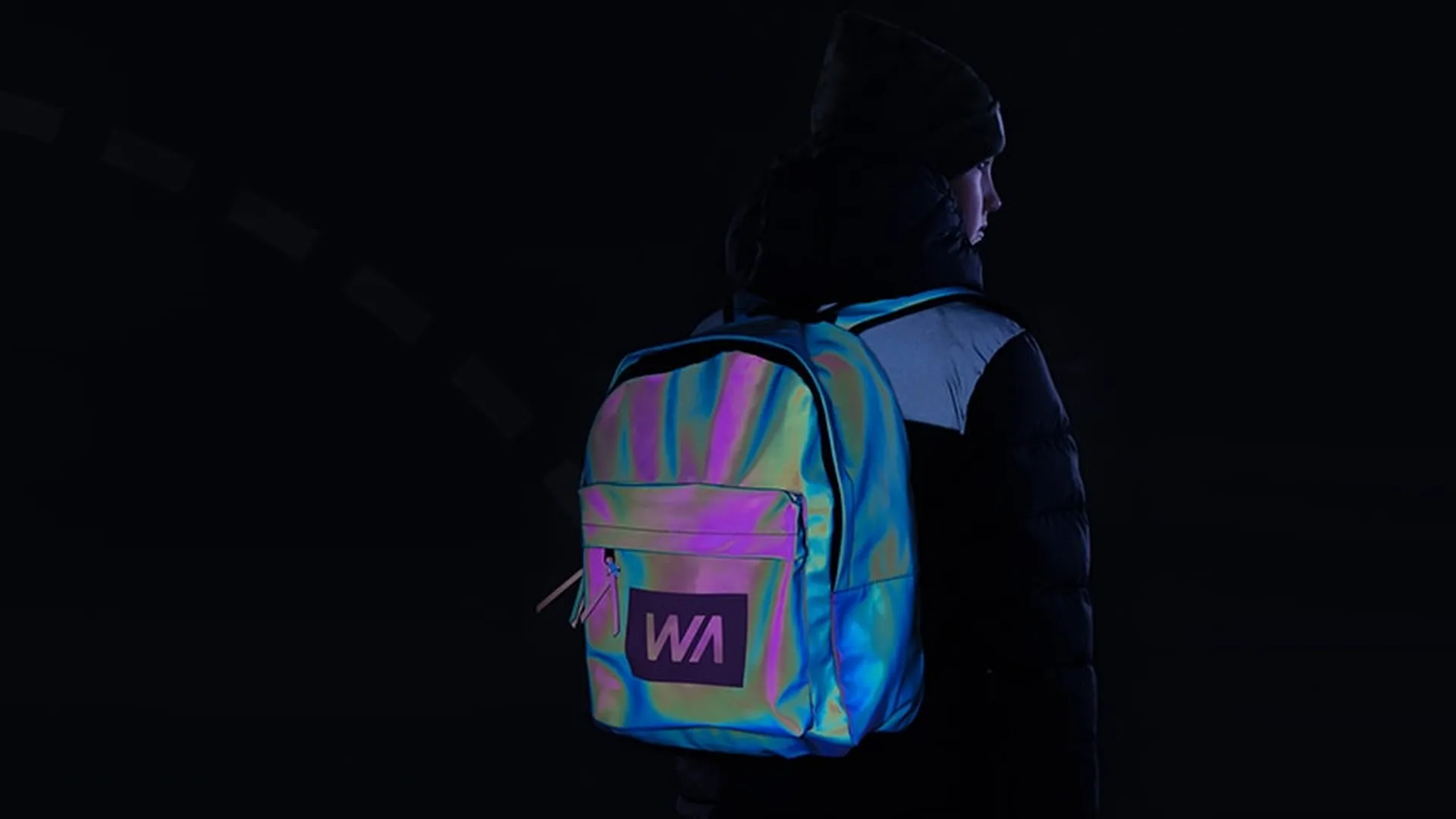 Back view of a person wearing a black jacket and beanie, carrying an iridescent, reflective backpack with a ‘WA’ logo, photographed against a dark background.