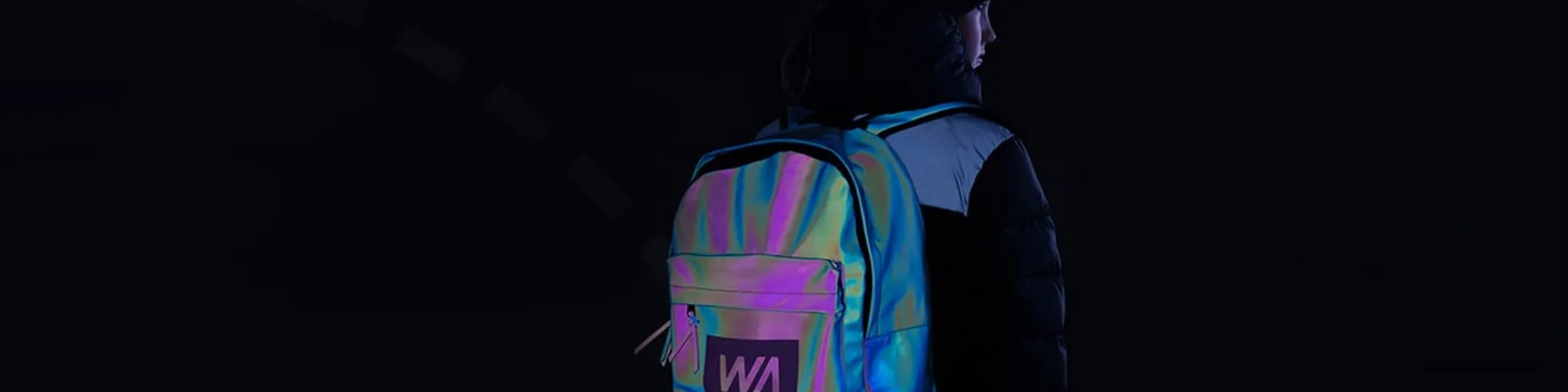 Back view of a person wearing a black jacket and beanie, carrying an iridescent, reflective backpack with a ‘WA’ logo, photographed against a dark background.