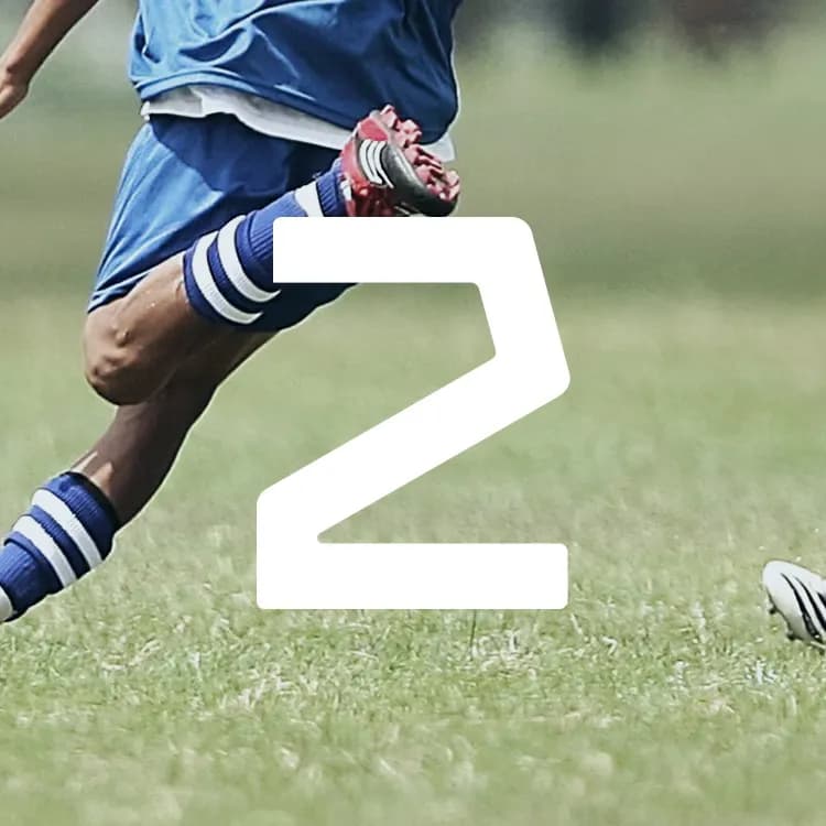 Soccer player in blue kicks the ball on a grassy field, wearing striped socks. Facing an opponent, a large number "2" overlays the image.