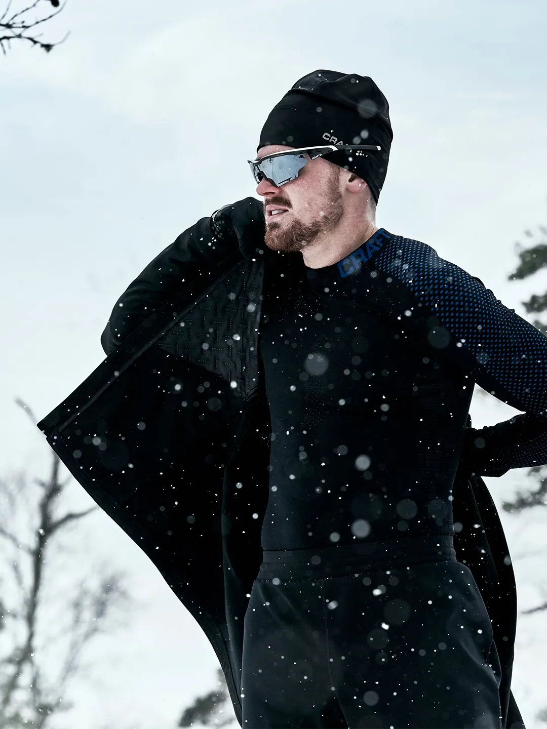 Man in winter athletic gear stands in snowy landscape, adjusting black jacket, wearing beanie and sunglasses, with snowflakes falling.
