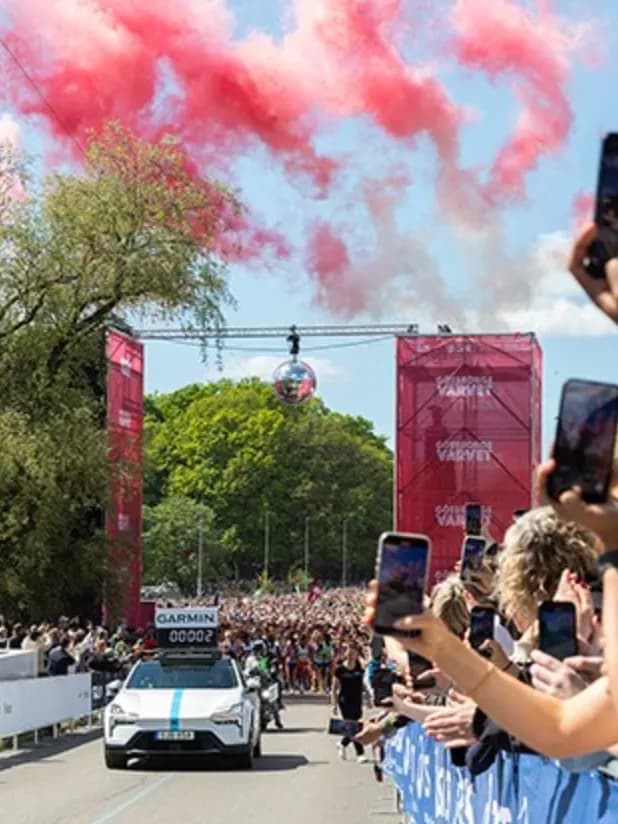 Crowd at a race, cheering and taking photos as a car crosses the finish line. Pink smoke fills the sky above the event entrance.