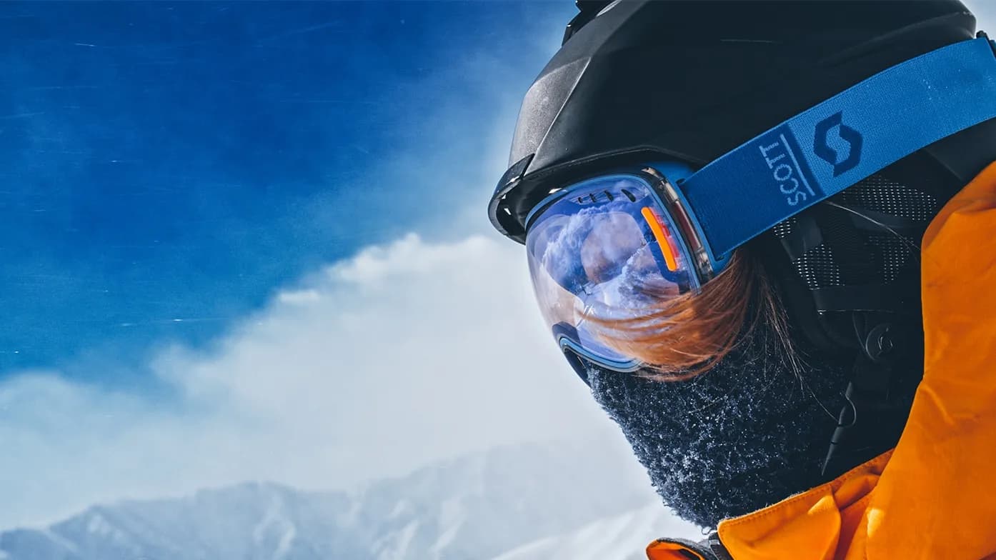 Person in orange jacket and ski goggles, facing snowy mountains under a clear blue sky, wearing a helmet and mask for cold weather protection.
