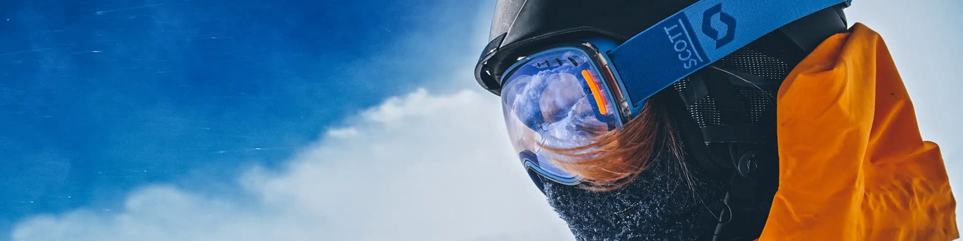 Person in orange jacket and ski goggles, facing snowy mountains under a clear blue sky, wearing a helmet and mask for cold weather protection.
