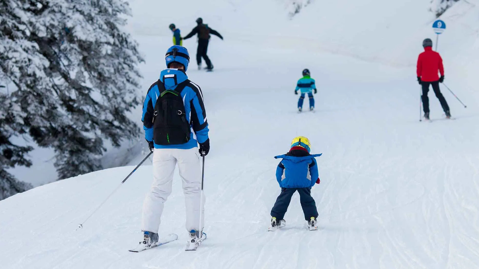 Skiers of varying ages and skill levels glide down a snowy slope, surrounded by snow-covered trees.