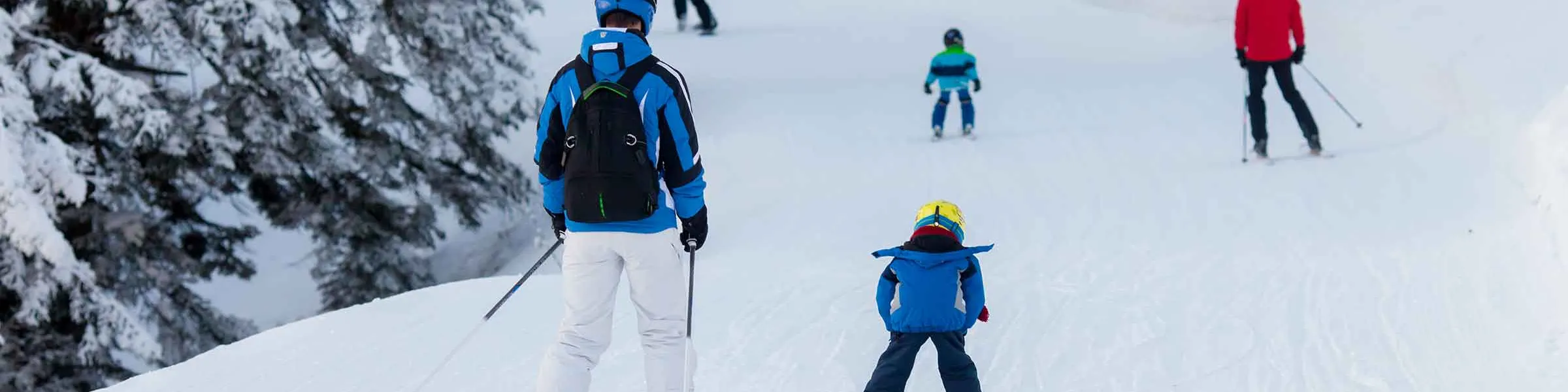 Skiers of varying ages and skill levels glide down a snowy slope, surrounded by snow-covered trees.