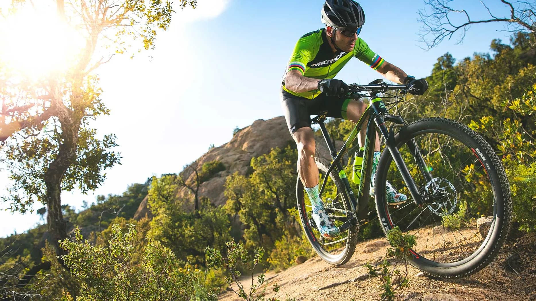 Cyclist in green jersey riding a mountain bike uphill on a rocky trail, sunlight filtering through trees in the background.