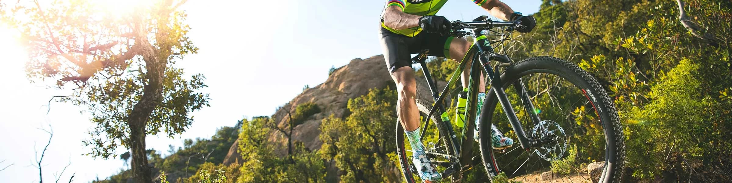 Cyclist in green jersey riding a mountain bike uphill on a rocky trail, sunlight filtering through trees in the background.