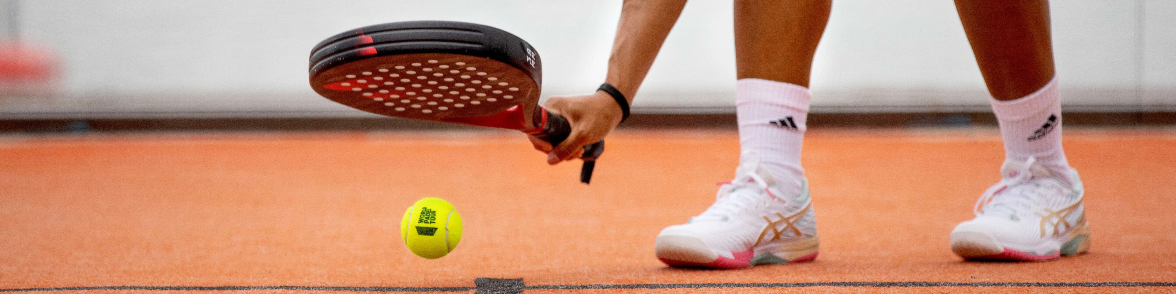 Person in white sportswear playing padel on an orange court, bending to hit a yellow ball with a black racket.