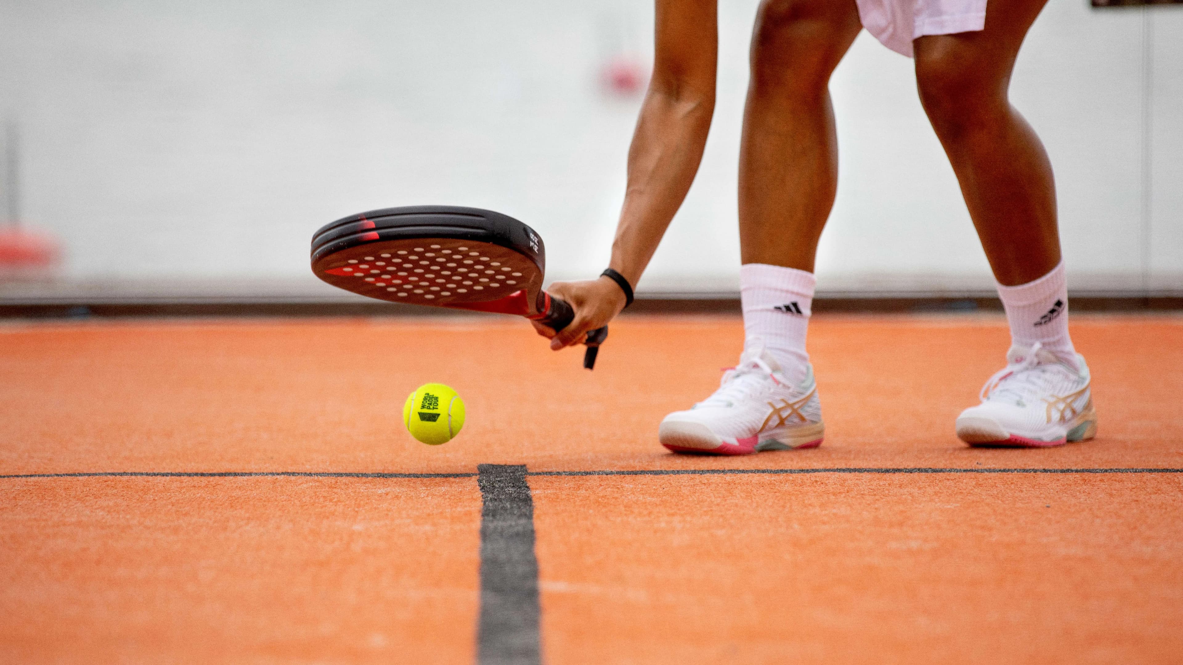 Person in white sportswear playing padel on an orange court, bending to hit a yellow ball with a black racket.