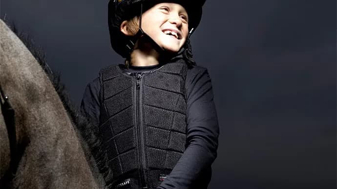 A happy child riding a horse, wearing a safety vest and a riding helmet.