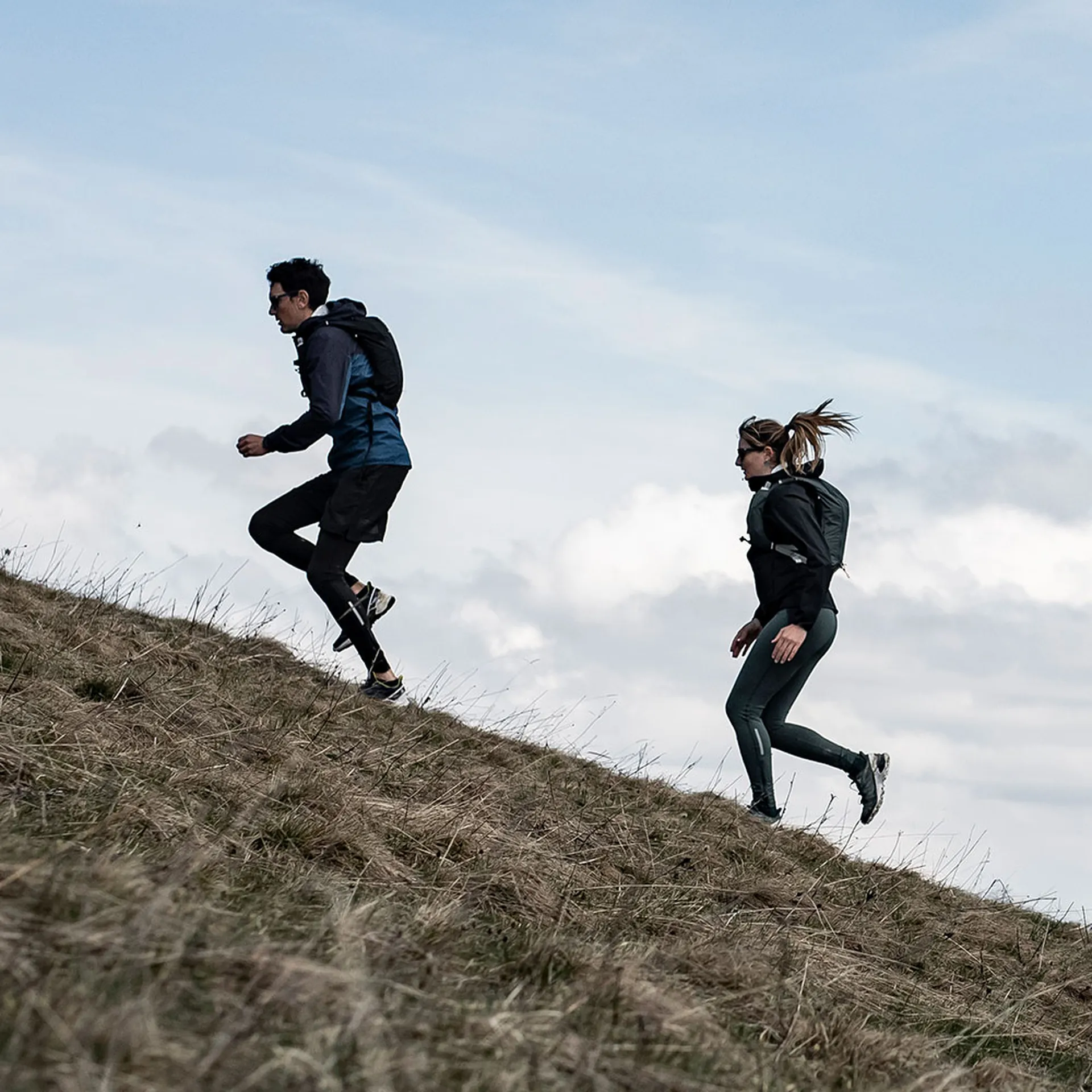 Two people trail running uphill on a grassy slope, wearing backpacks and athletic clothing, with an open sky in the background