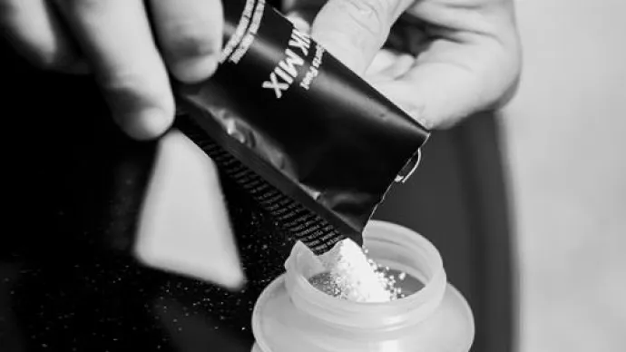 A hand pours protein powder from a black packet into a plastic shaker bottle, in black and white.